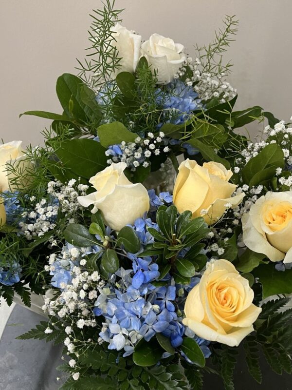 A table floral arrangement with pastel yellow roses, blue hydrangeas, white baby's breath and greenery